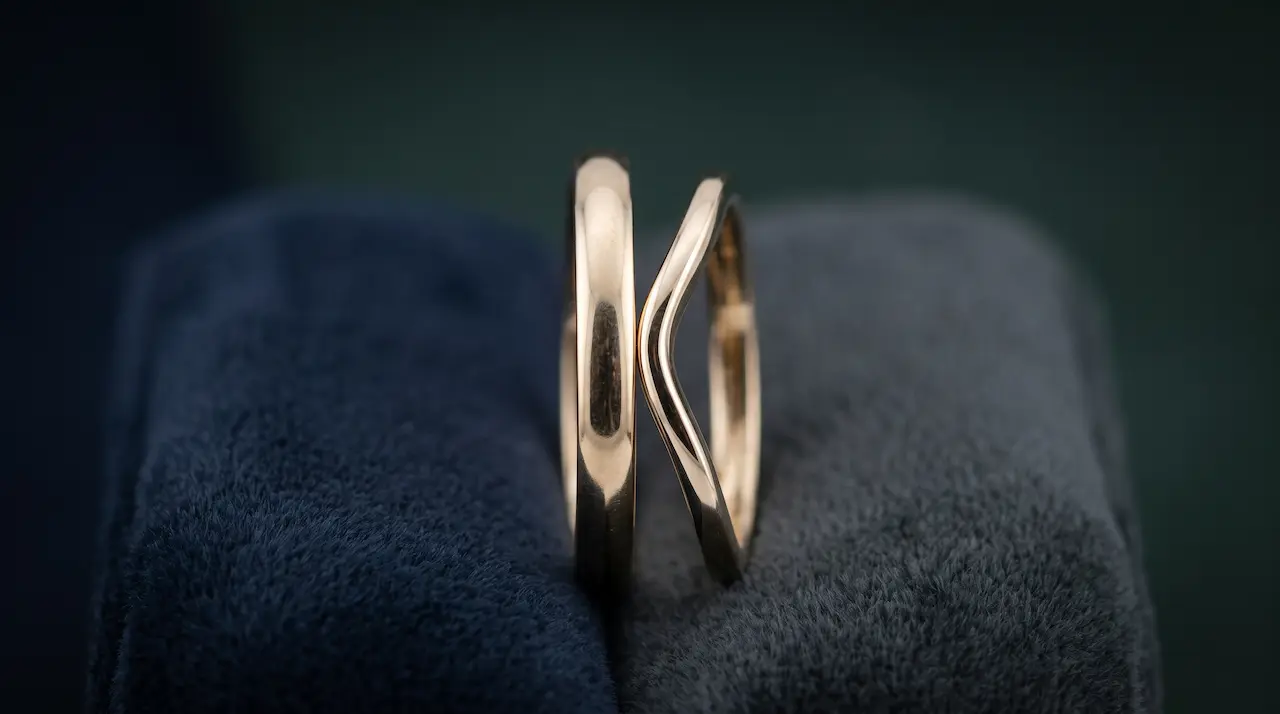 Photo professionnelle de deux alliances de profil posées côte à côte sur un présentoir en velours, l'une droite et l'une échancrée, mise en évidence du contact entre les anneaux, éclairage de studio doux, style macro épuré, format 16:9.