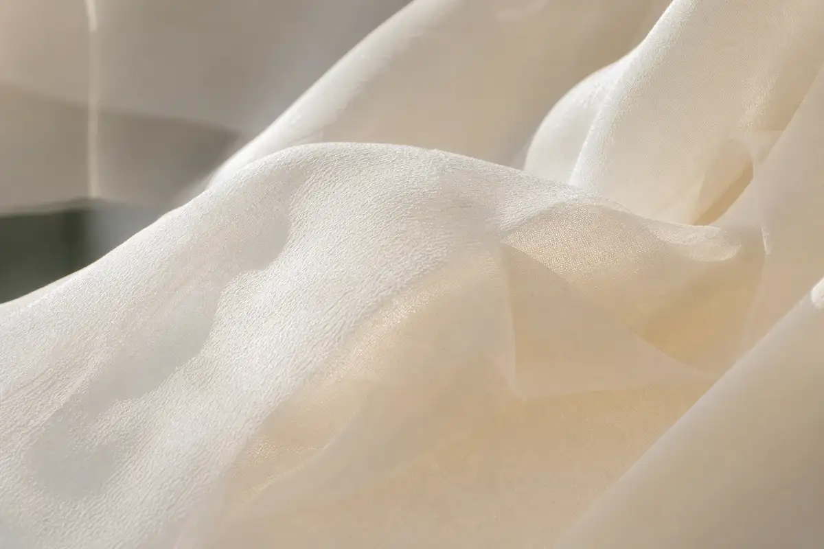 Photo professionnelle en gros plan d'un tissu de robe de mariée en mousseline de soie ivoire légèrement froissé par la brise, texture détaillée visible, lumière naturelle latérale, style éditorial minimaliste, format 16:9
