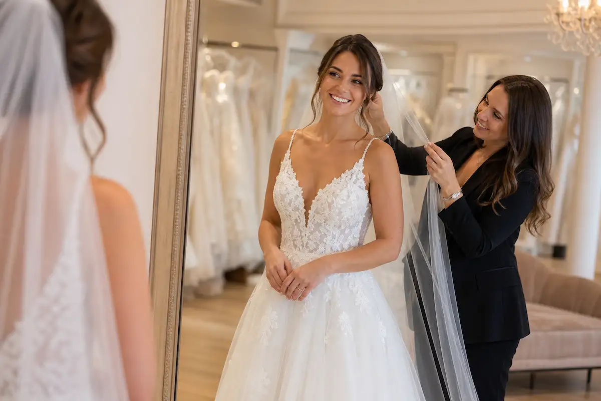Photo professionnelle d'un essayage de robe de mariée dans une boutique lumineuse, mariée souriante devant un grand miroir, conseillère aux côtés ajustant le voile, robes en exposition en arrière-plan flou, éclairage chaleureux intérieur, style naturel et bienveillant, format 16:9