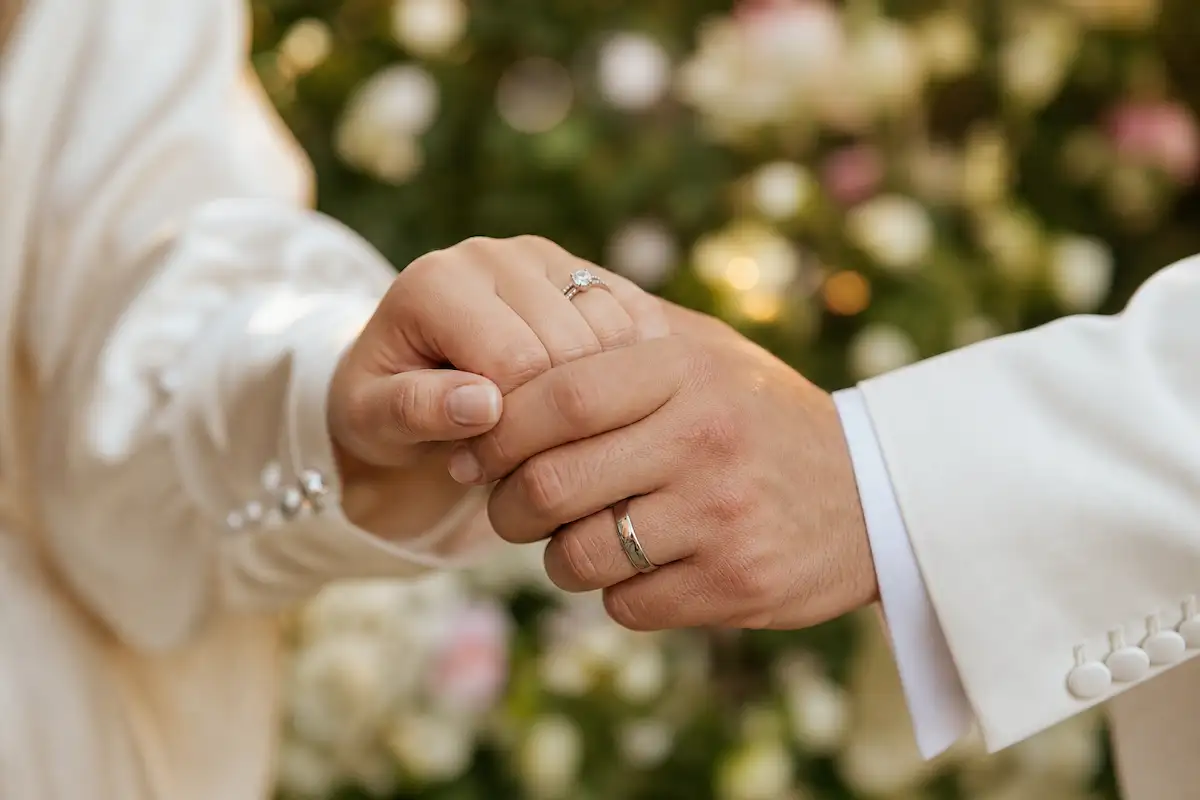 Photo professionnelle en gros plan de deux mains enlacées, alliance visible, robe de mariée en soie ivoire et manchette de costume blanche au premier plan, fond jardin flou en bokeh, éclairage naturel doux, style romantique, format 16:9