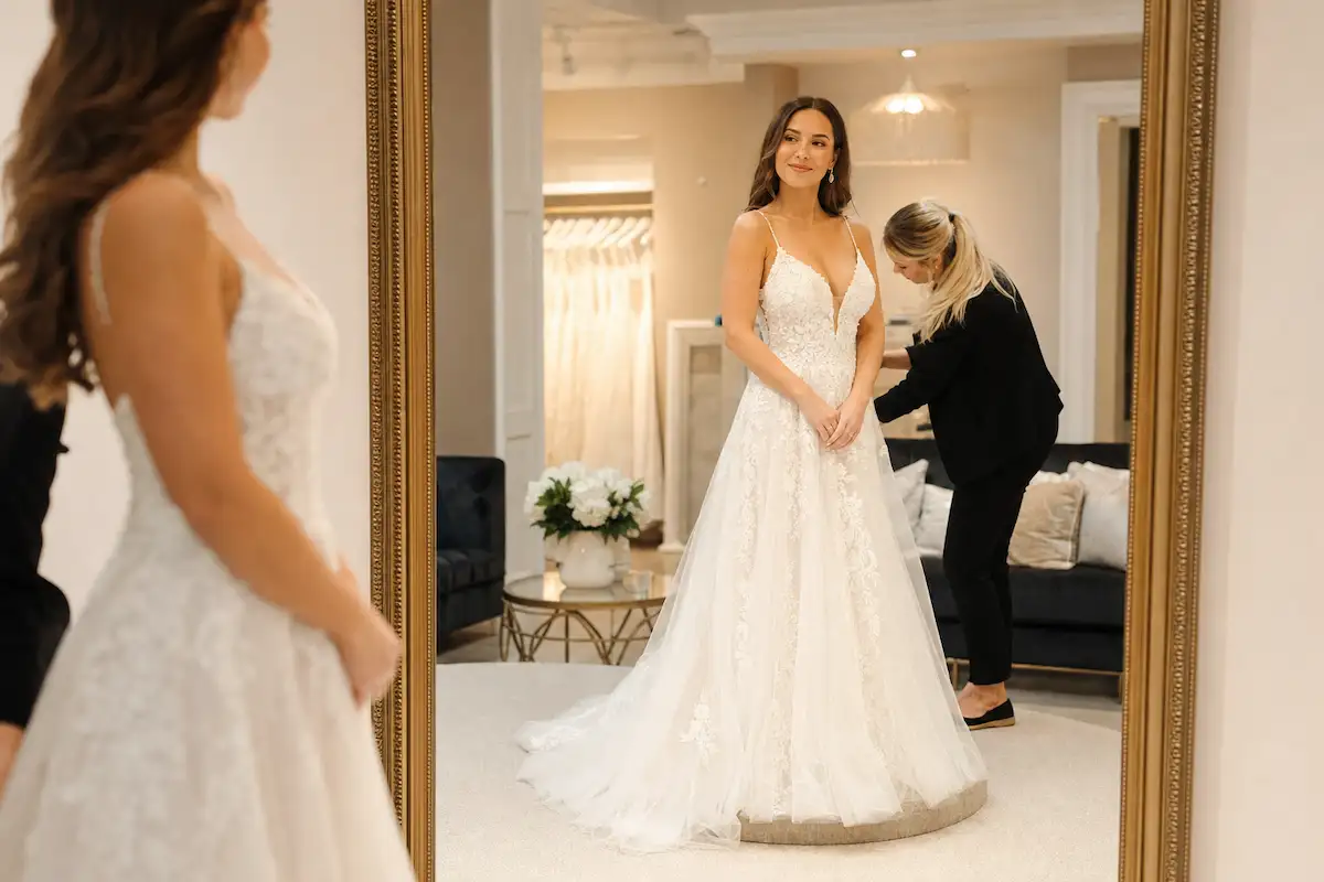 Photo professionnelle d'une mariée en robe longue blanche essayant différentes postures devant un grand miroir en boutique, conseillère en arrière-plan qui ajuste un détail, expression de satisfaction naturelle, éclairage chaleureux, style bienveillant et professionnel, format 16:9